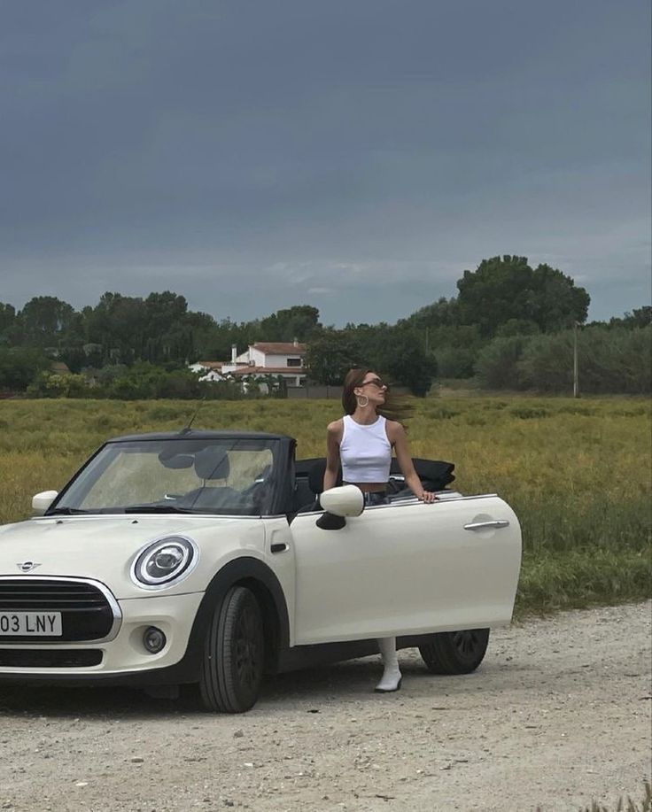 a woman sitting in the driver's seat of a white convertible car on a dirt road