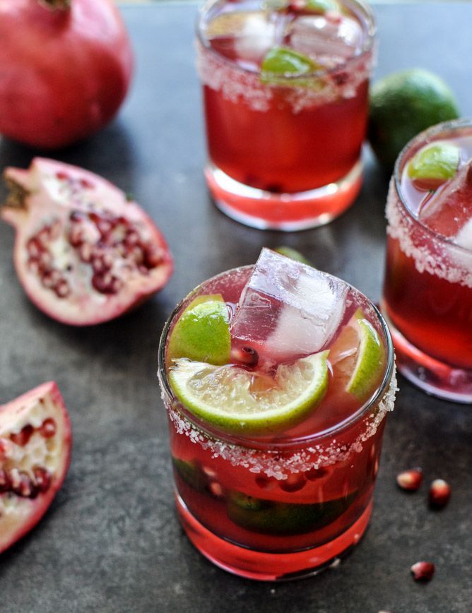 two glasses filled with drinks and garnished with limes next to pomegranates