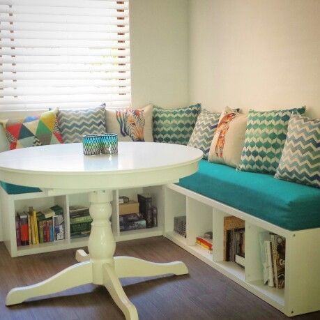 a white table sitting under a window next to a book shelf filled with books and pillows