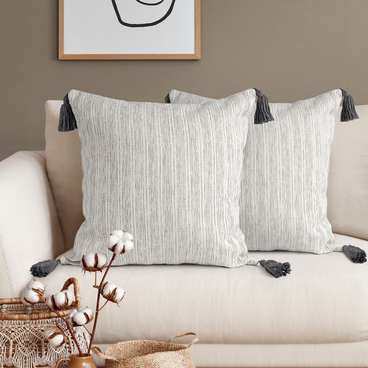 two pillows on a white couch in front of a painting and basket with cotton flowers