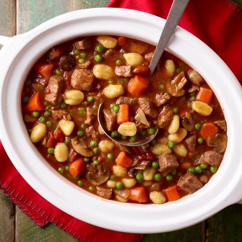 a white bowl filled with meat and vegetables on top of a red cloth next to a spoon