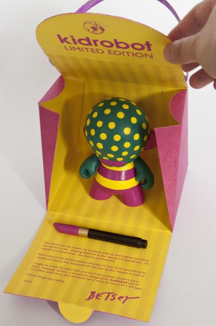 a hand is holding a pen in a box with a doll inside it and the lid open