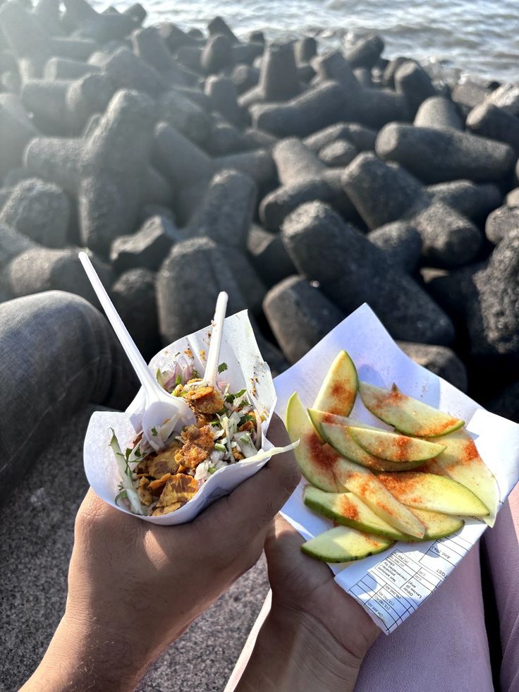 a person holding up a paper container filled with food next to some rocks and water