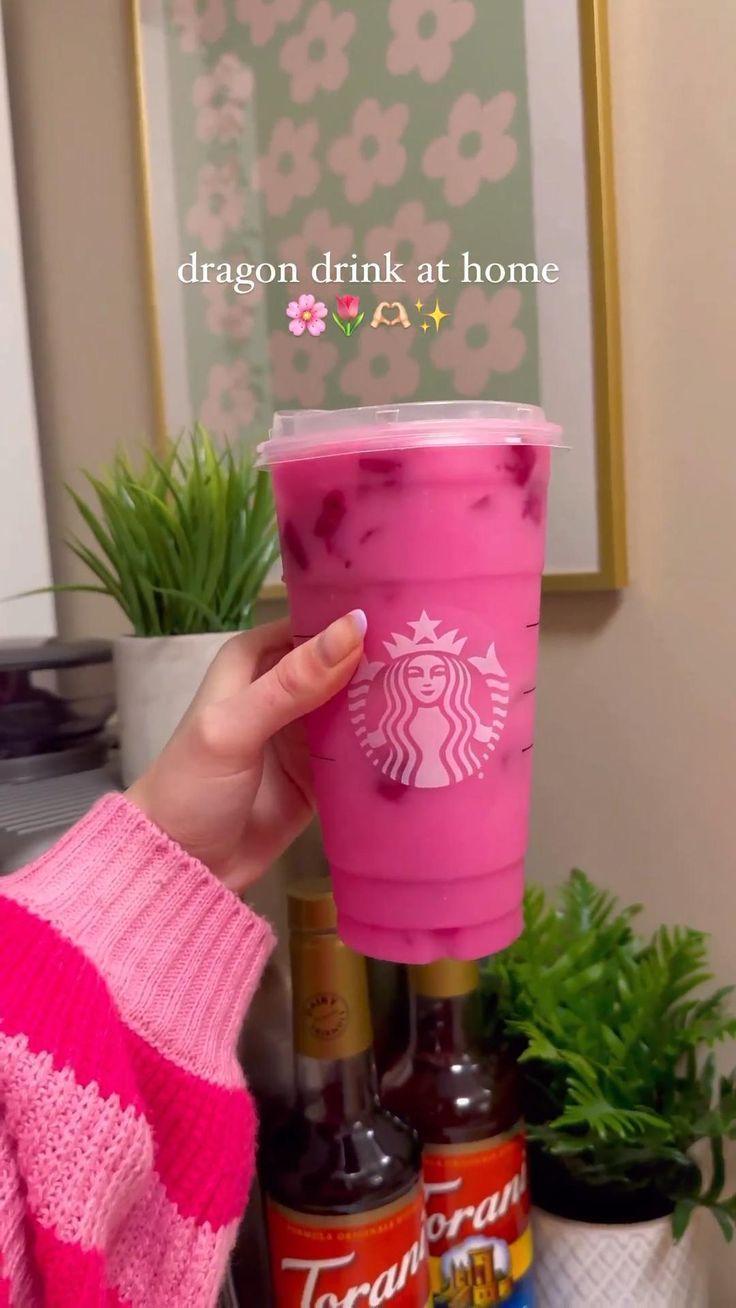 a woman holding up a pink starbucks cup in front of some plants and other items