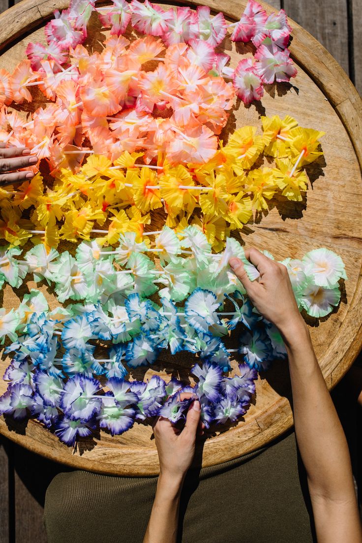 two hands reaching for colorful flowers on a wooden tray with string in the middle and one hand holding onto it