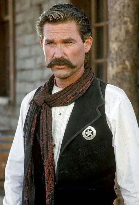 a man with a moustache wearing a black vest and brown scarf around his neck