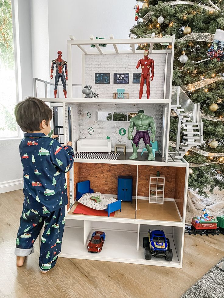 a young boy playing with a toy house in front of a christmas tree and spiderman figurines