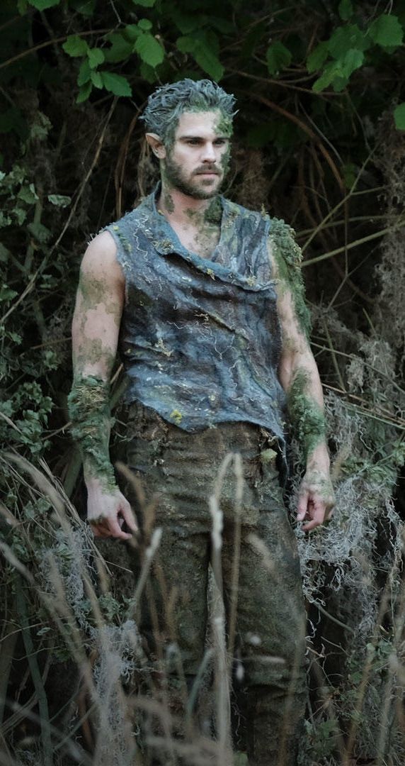 a man with green paint on his face standing in tall grass and bushes, looking at the camera