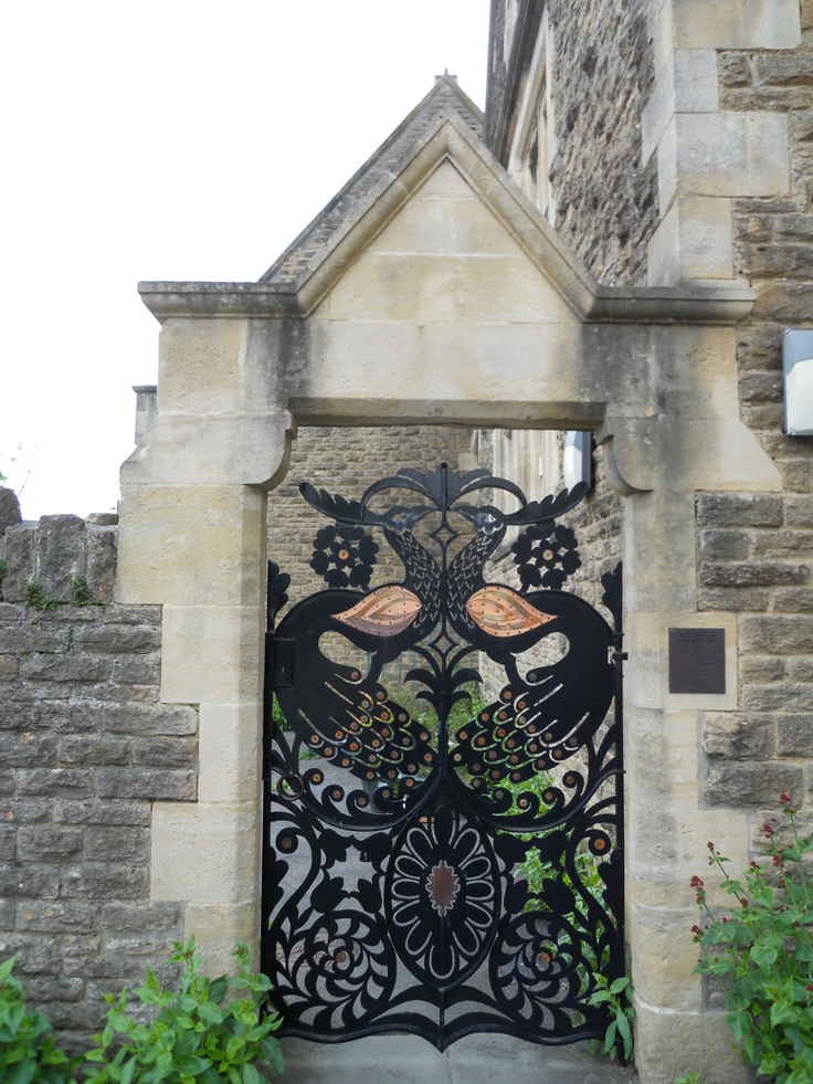 an iron gate with flowers and birds on it in front of a stone building that is surrounded by greenery