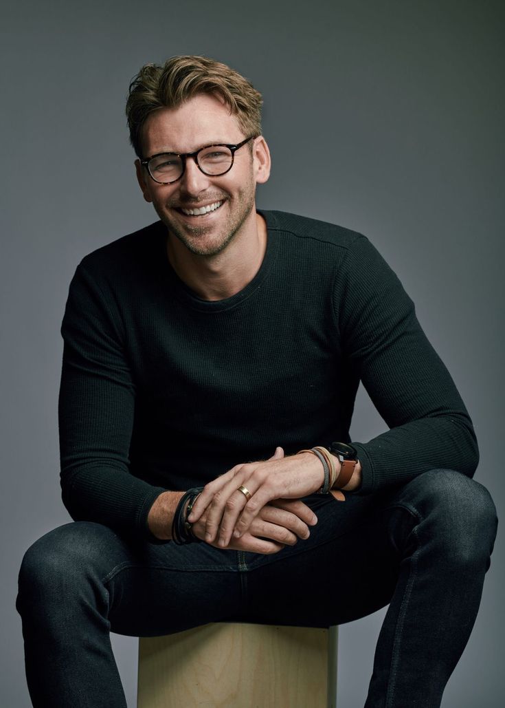 a man with glasses sitting on top of a wooden stool smiling at the camera while wearing a black sweater and jeans