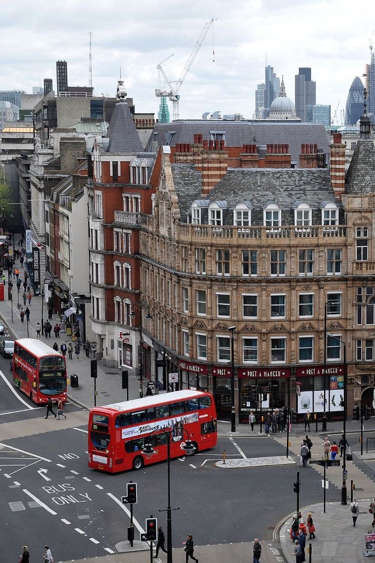 two red double decker buses driving down a street next to tall buildings in the city