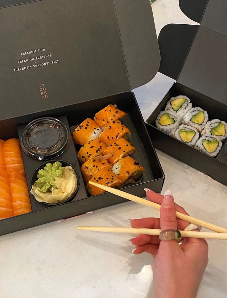 a person holding chopsticks in front of a box of sushi and vegetables