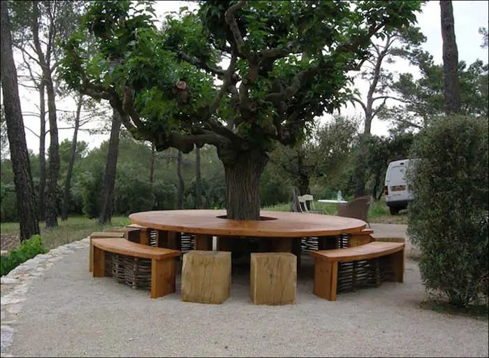 a large wooden table sitting under a tree in the middle of a park with benches underneath it