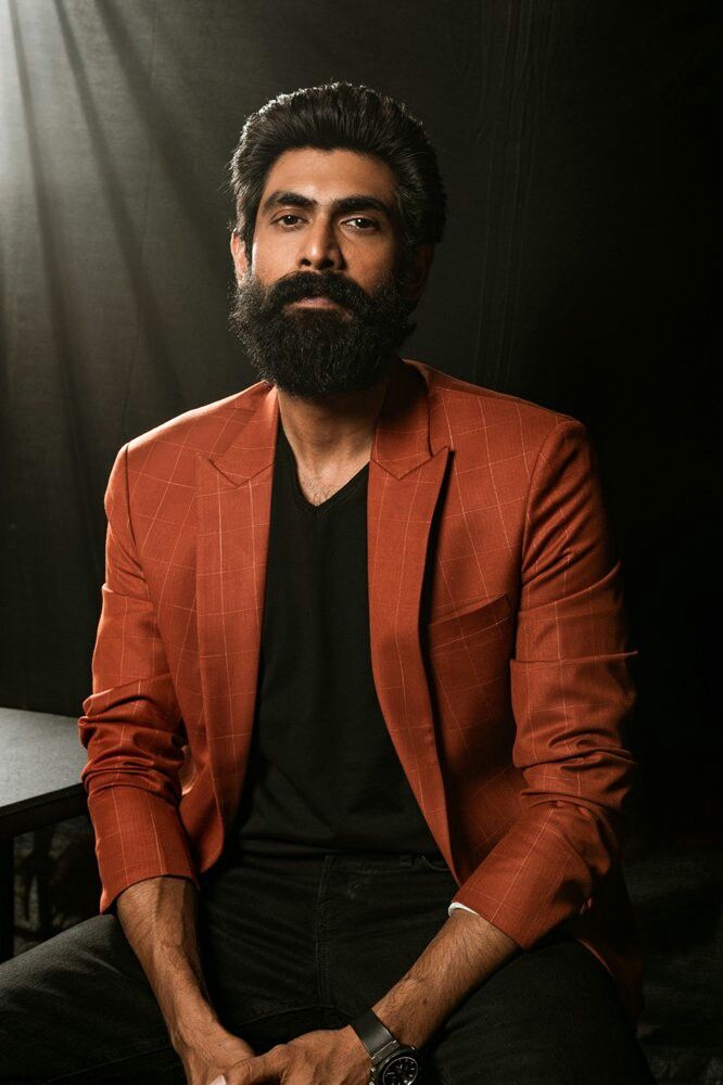 a man with a beard sitting in front of a black background wearing a red blazer