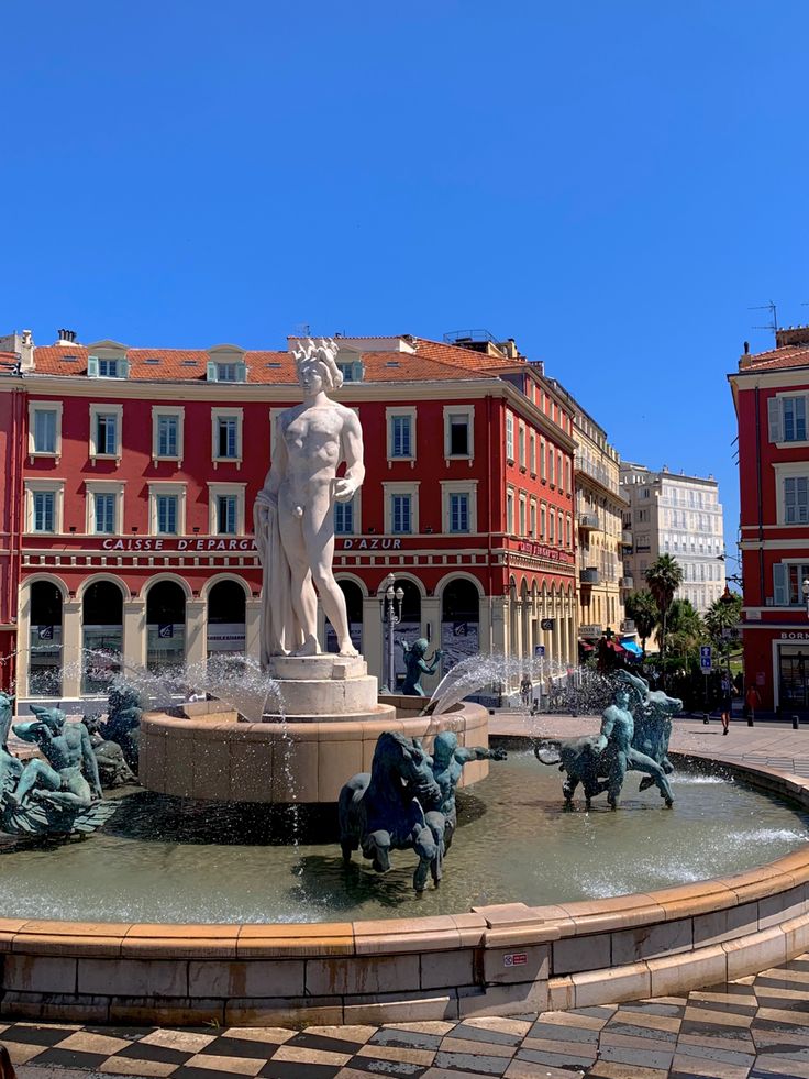 a fountain in front of a red building with statues on it and people walking around