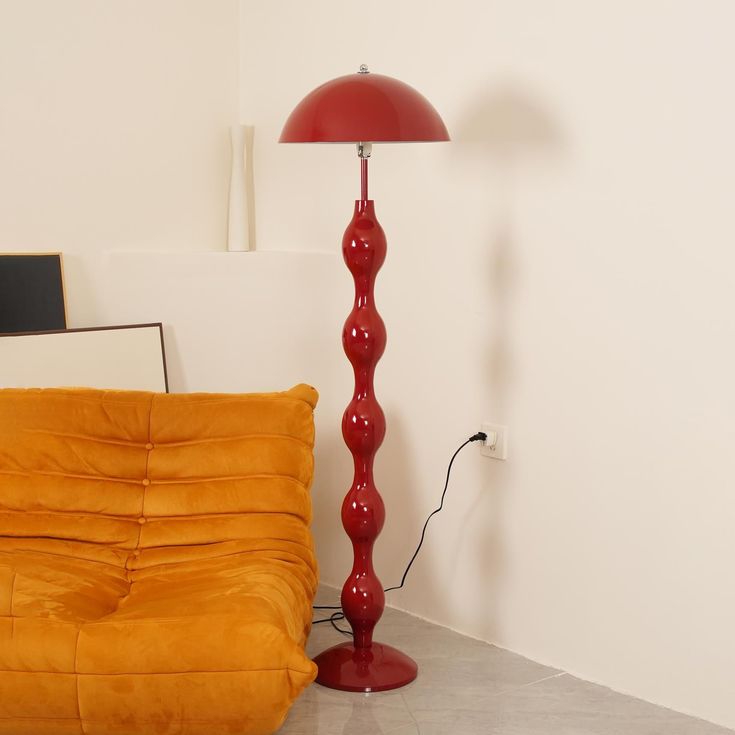 a red floor lamp sitting next to a yellow couch in a room with white walls