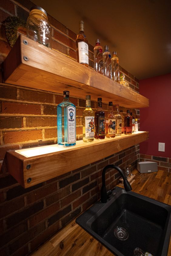 a kitchen sink sitting under a wooden shelf next to bottles on top of a brick wall