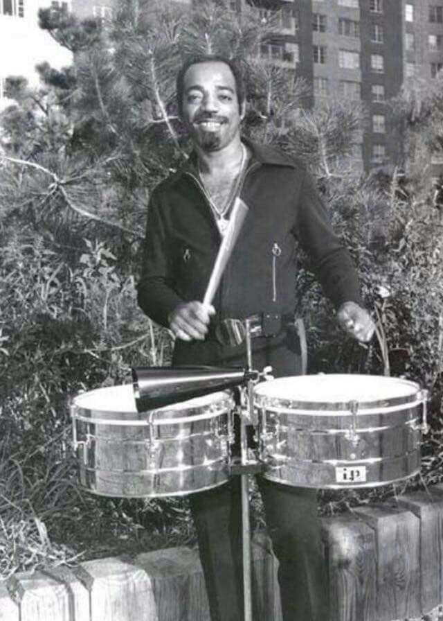 a man is playing the drums in front of some trees and bushes with buildings behind him