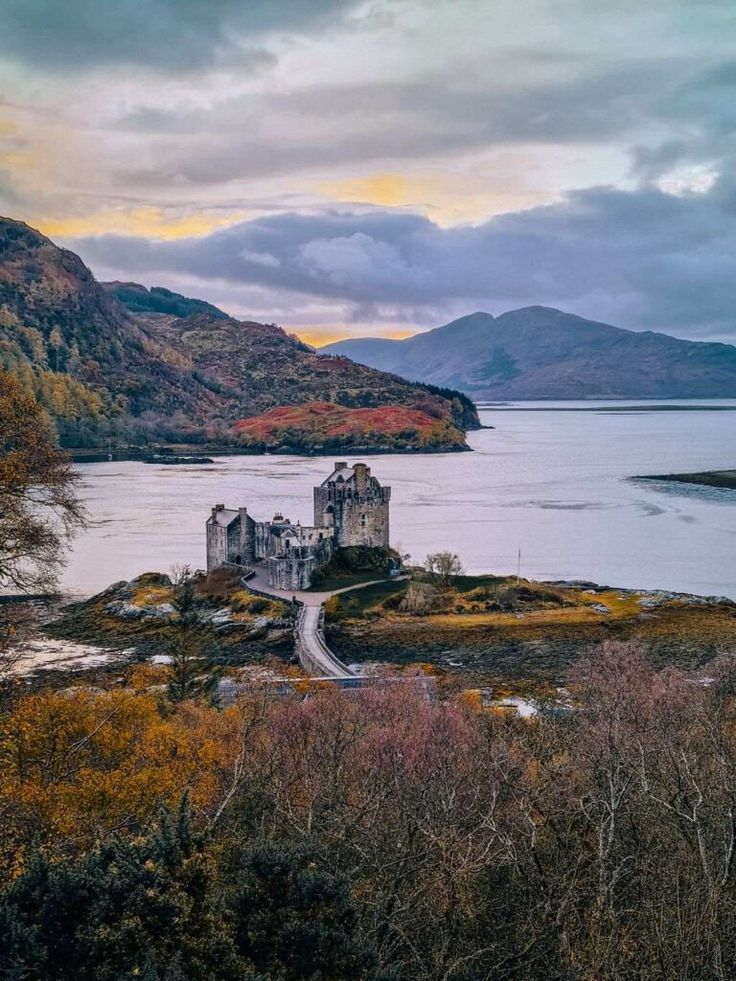 an old castle sitting on top of a hill next to a body of water with mountains in the background