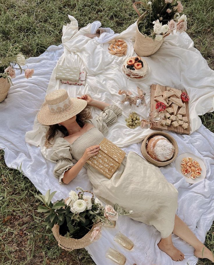 a woman laying on top of a white blanket next to food and drinks in baskets