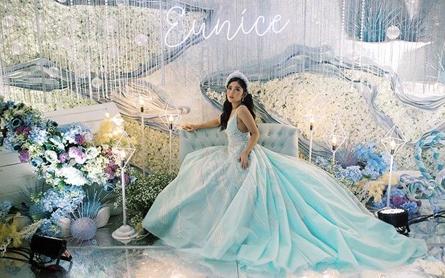 a woman in a blue dress sitting on a couch with flowers and chandelier