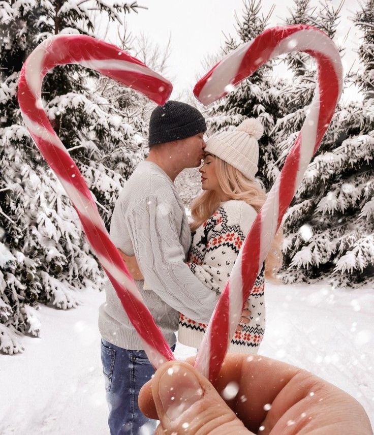 a man and woman holding candy canes in front of snow covered pine trees with heart shaped lollipops