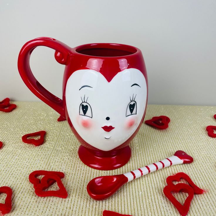 a red coffee mug with a face painted on the side and spoons scattered around it