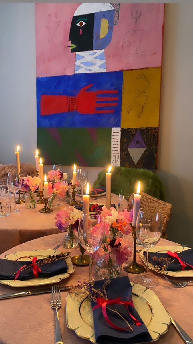 the table is set with place settings and candles in front of a colorful painting on the wall