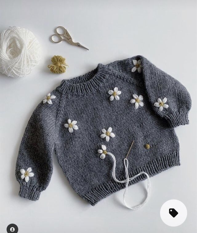 a gray sweater with white flowers on it next to yarn and crochet hooks