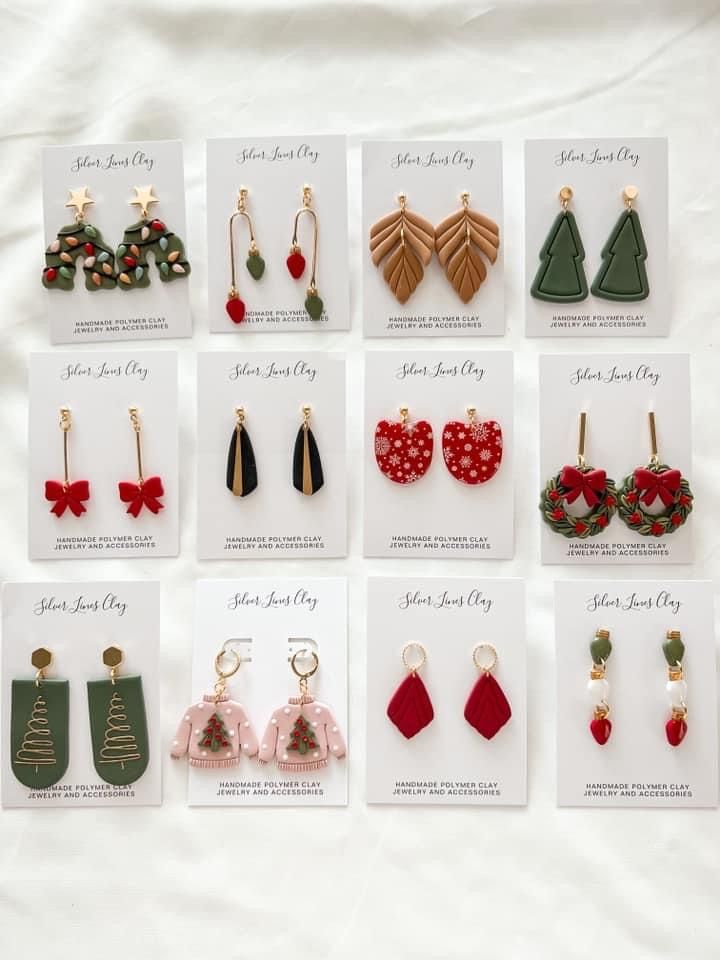 twelve pairs of christmas earrings are displayed on a white sheet, with holiday cards attached to them