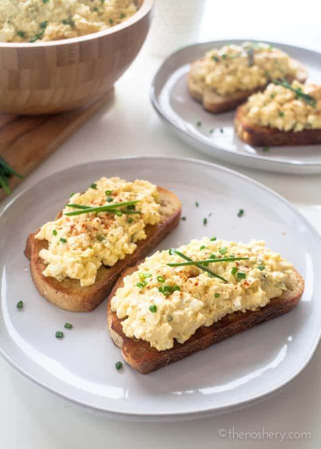 two white plates topped with toasted bread covered in scrambled eggs next to a bowl of chives