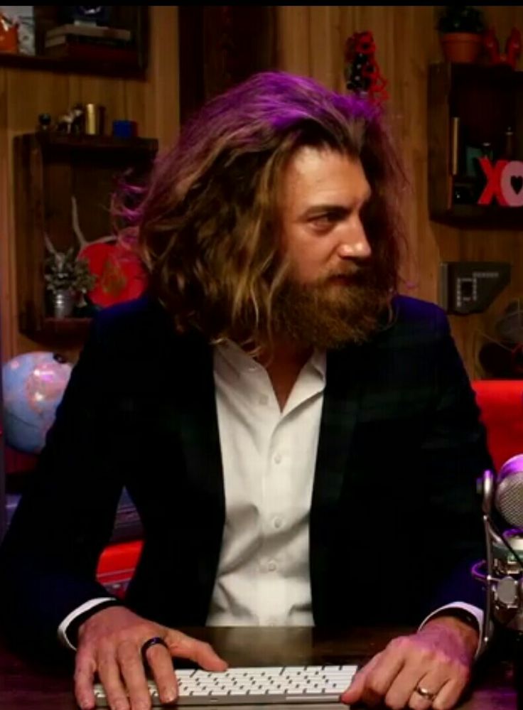 a man with long hair sitting at a desk in front of a keyboard and microphone