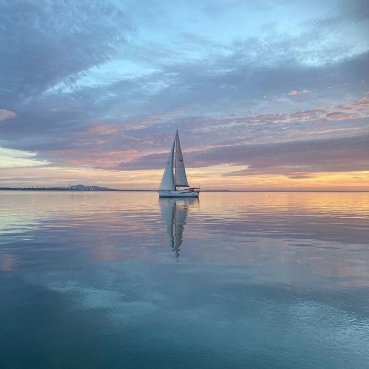 a sailboat floating on top of a large body of water at sunset with clouds in the sky