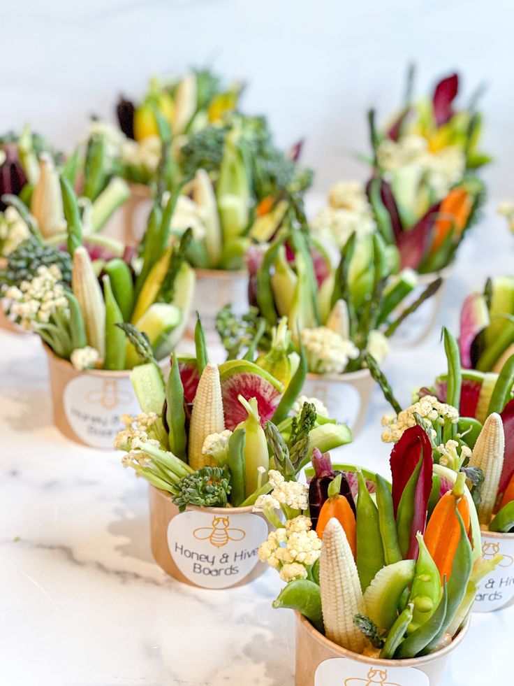 there are many small cups with flowers in them on the white tablecloth and one is filled with broccoli, carrots, celery, corn, and other vegetables