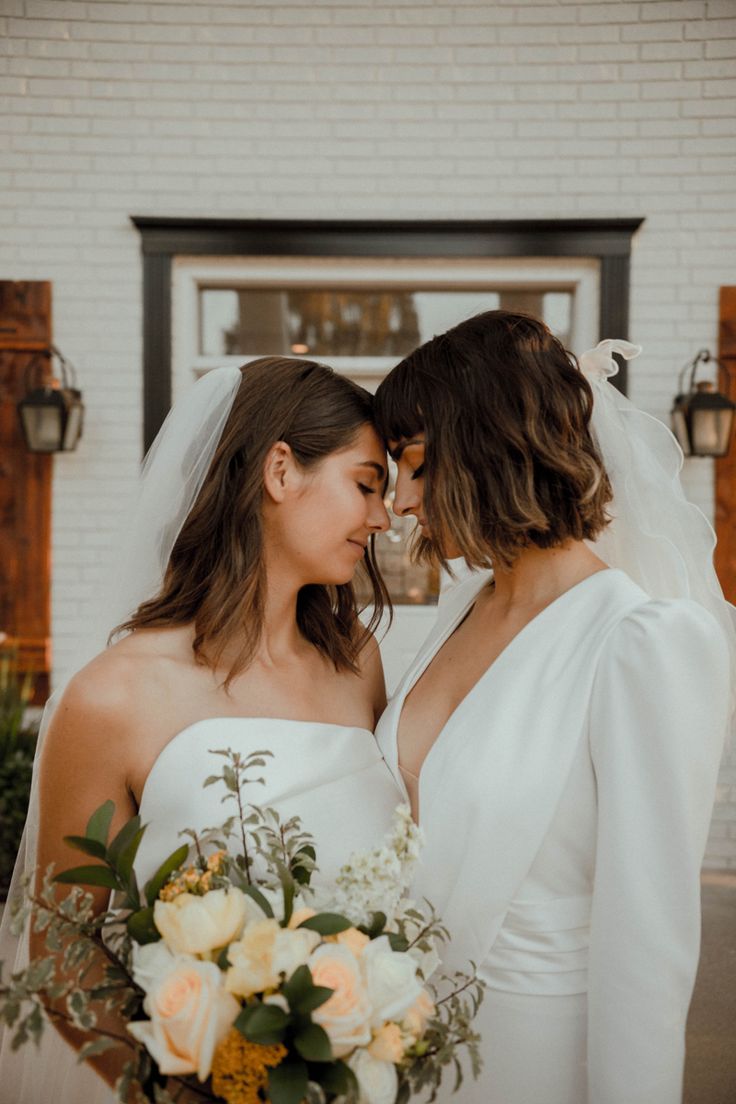 two women in white dresses standing next to each other holding bouquets and looking at each other