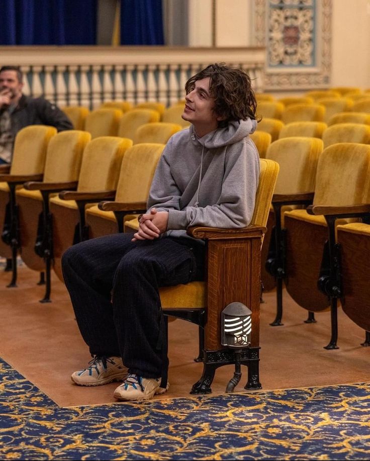 a man sitting on a chair in front of rows of chairs with microphones behind him