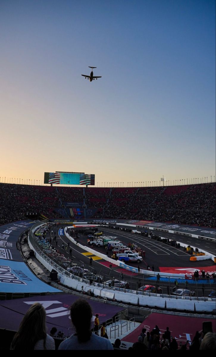 an air plane flying over a race track at sunset or dawn with spectators watching from the stands