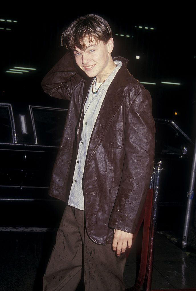 a young man standing in front of a car wearing brown pants and a leather jacket