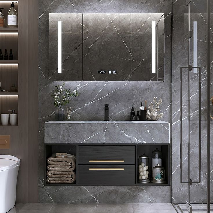 a bathroom with grey marble walls and flooring, lighted by lights above the sink