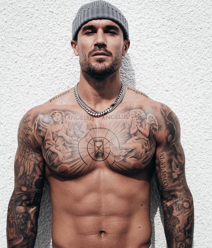 a shirtless man with tattoos on his chest and hands behind his back, standing in front of a white wall