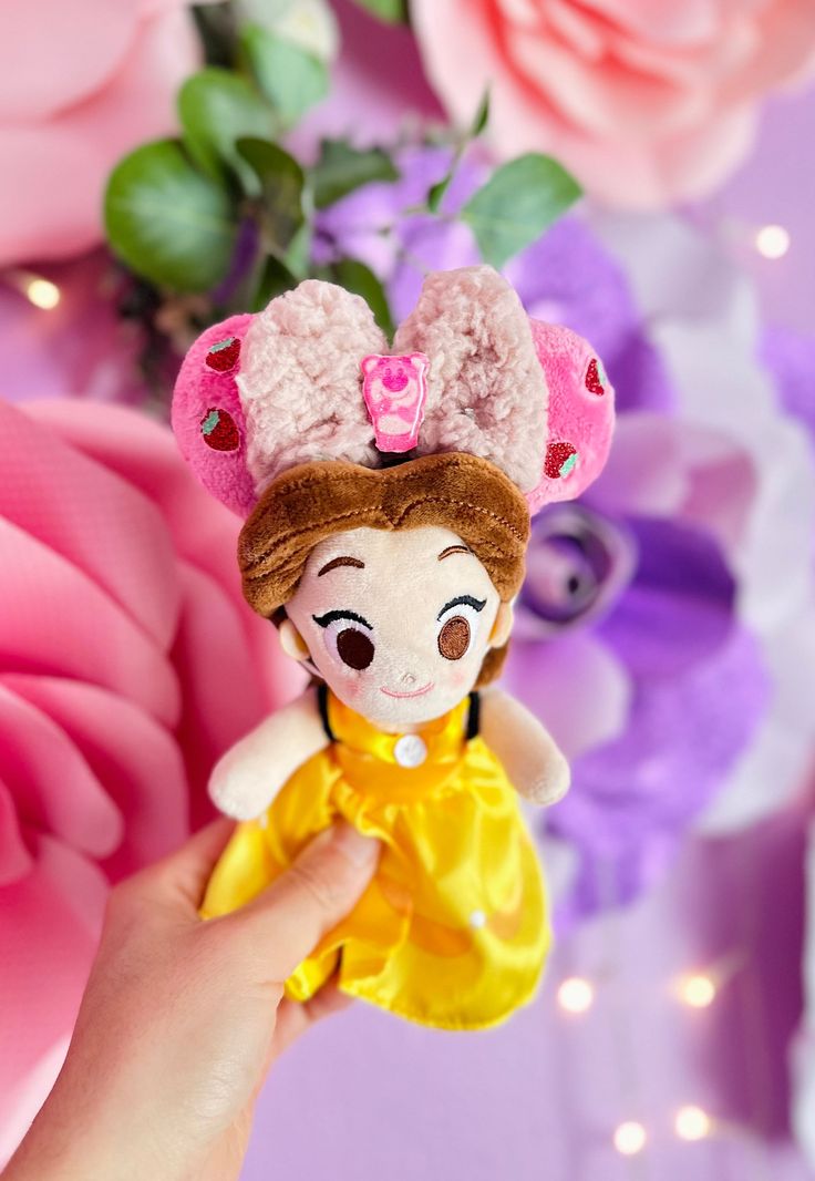 a hand holding a small doll in front of pink flowers