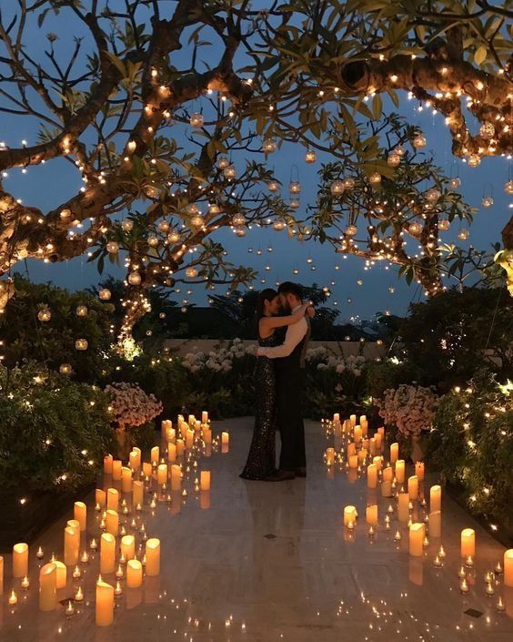 a couple is hugging in front of many lit candles and trees with lights all around them