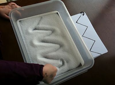 a person sitting at a table with a tray filled with sand and writing on it