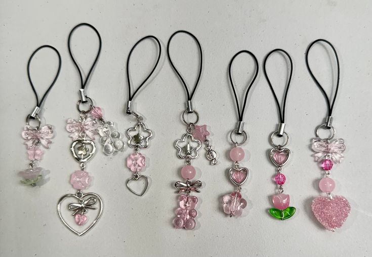 several charms are hanging on a white surface with black cords and pink beads, hearts, and bows