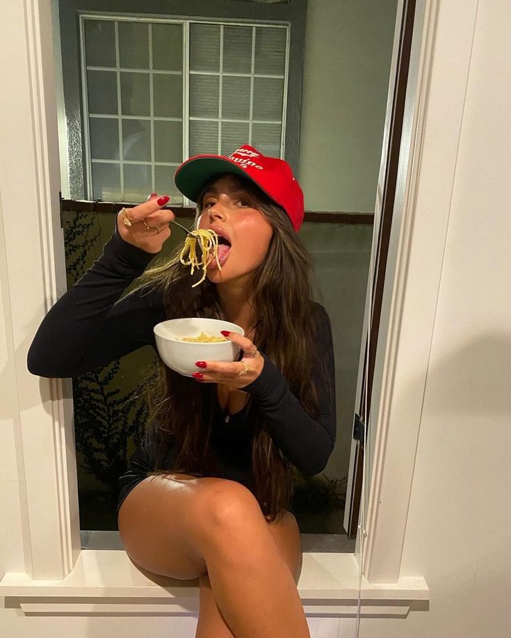 a woman is eating noodles out of a bowl while sitting on the toilet in front of a window