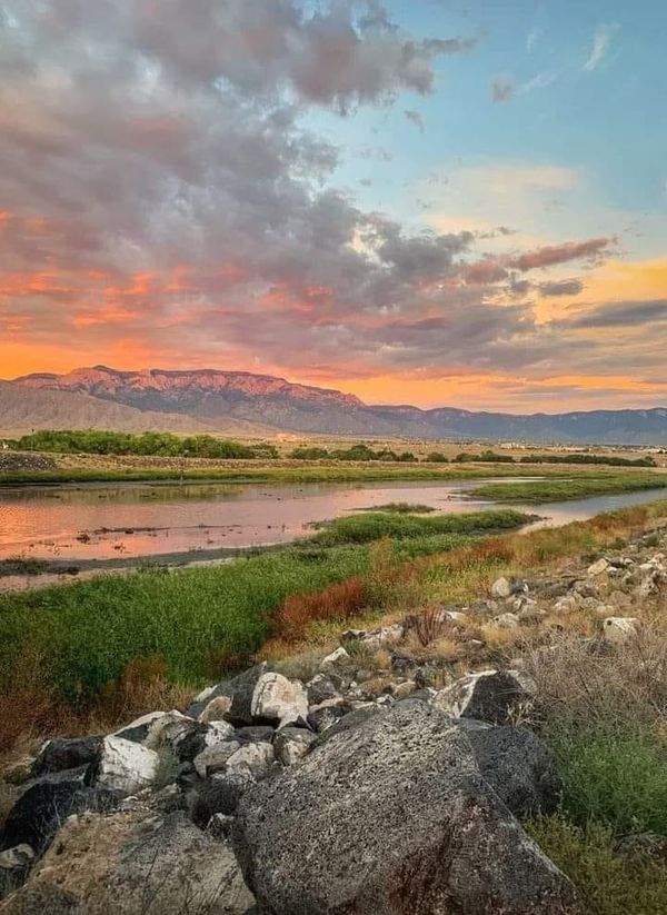 the sun is setting over a river with rocks and grass on both sides, and mountains in the distance