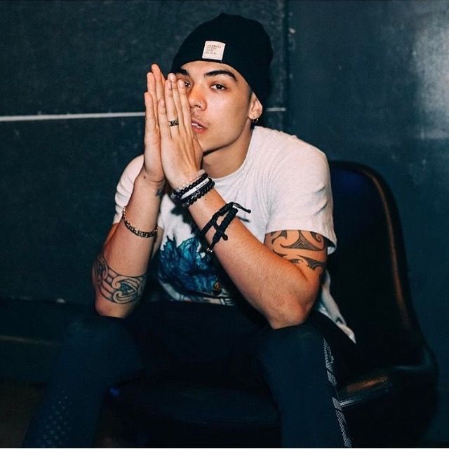 a young man sitting in a chair with his hands clasped up to his face while wearing a hat