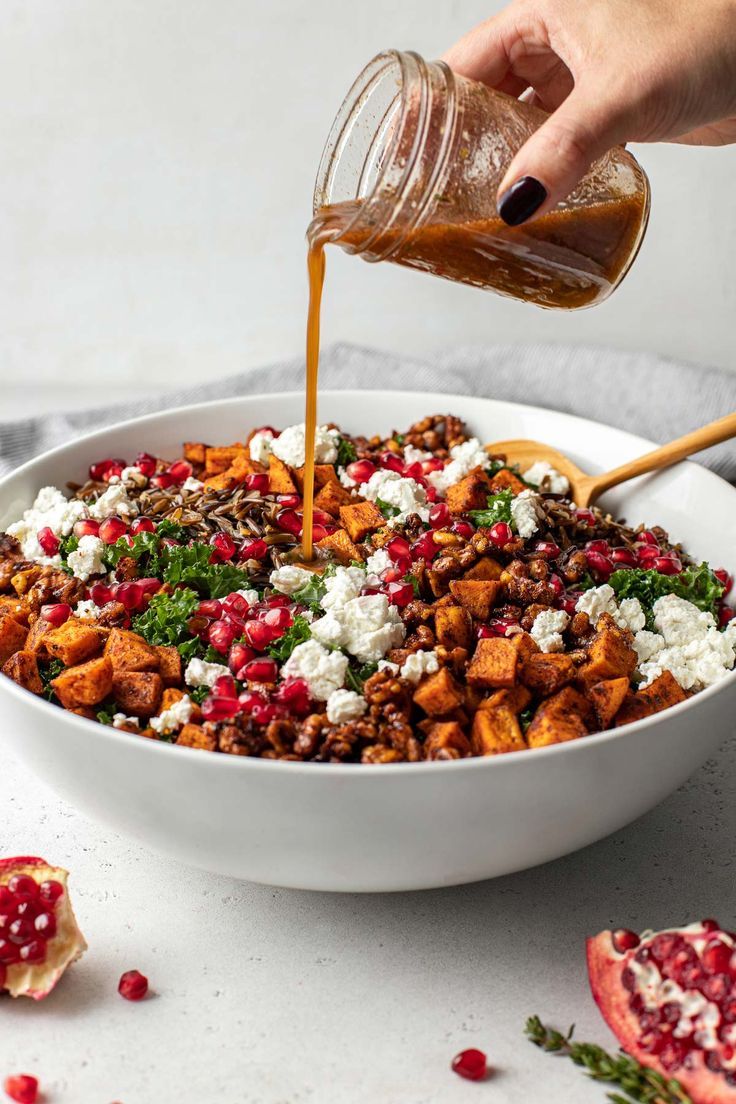 someone pouring dressing into a white bowl filled with food and garnished with pomegranates