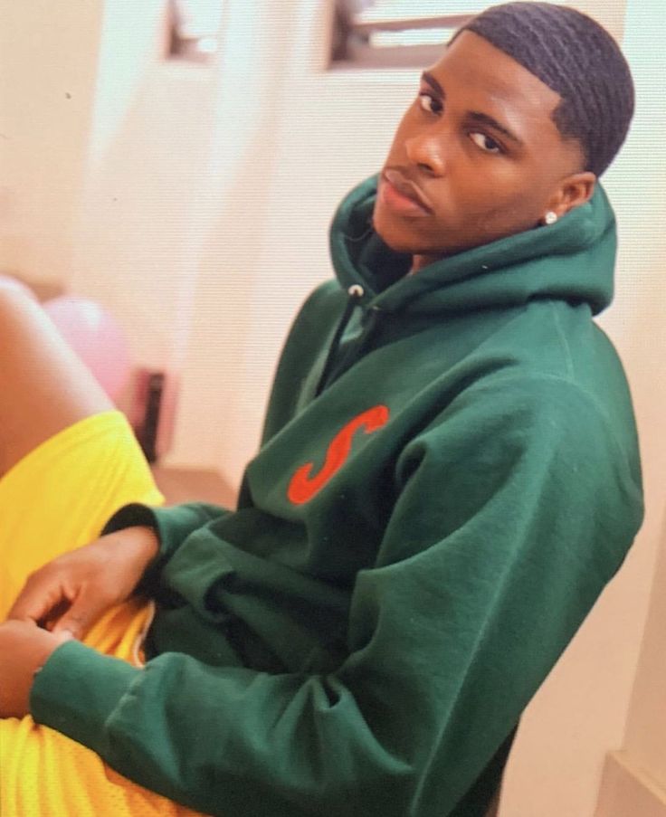 a man in a green hoodie and yellow skirt sitting on the floor with his legs crossed