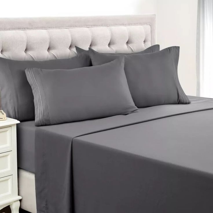 a bed with grey sheets and pillows in a bedroom next to a night stand on a white nightstand